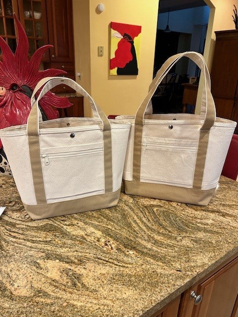 Two beige Totes for Everyday Use by Semen Tanks - Select Genetics, with front zippered pockets and 18 or 24 inch straps, rest on a granite kitchen countertop. A red rooster figurine and colorful artwork are in the background.