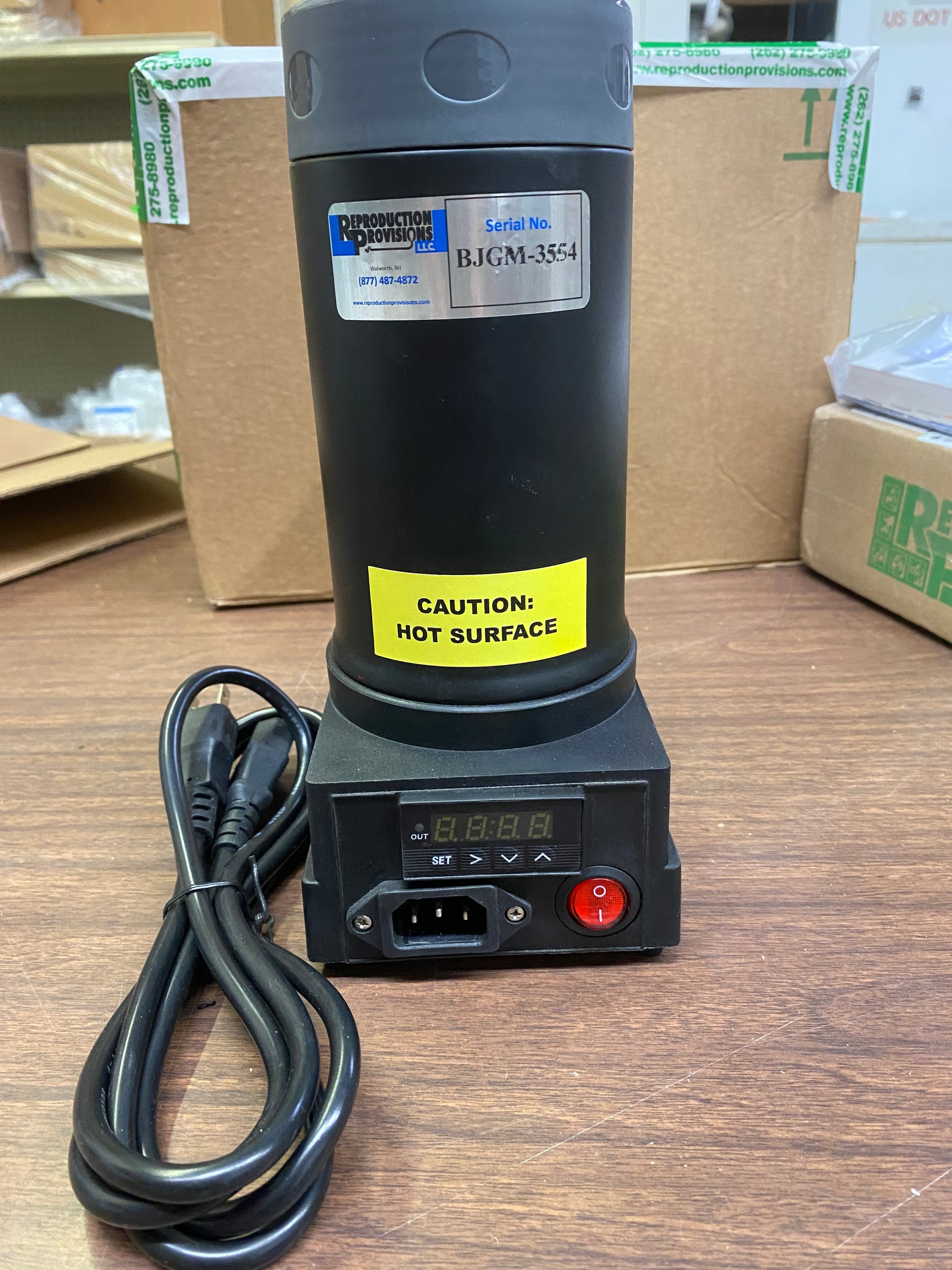 A black Digital Semen Thawing/Embryo Thawing Unit - 220V AC (1023024) by Semen Tanks - Select Genetics, labeled Caution: Hot Surface and Serial No. BJGM-3554, sits on a wooden table with display and power cord, boxes behind.