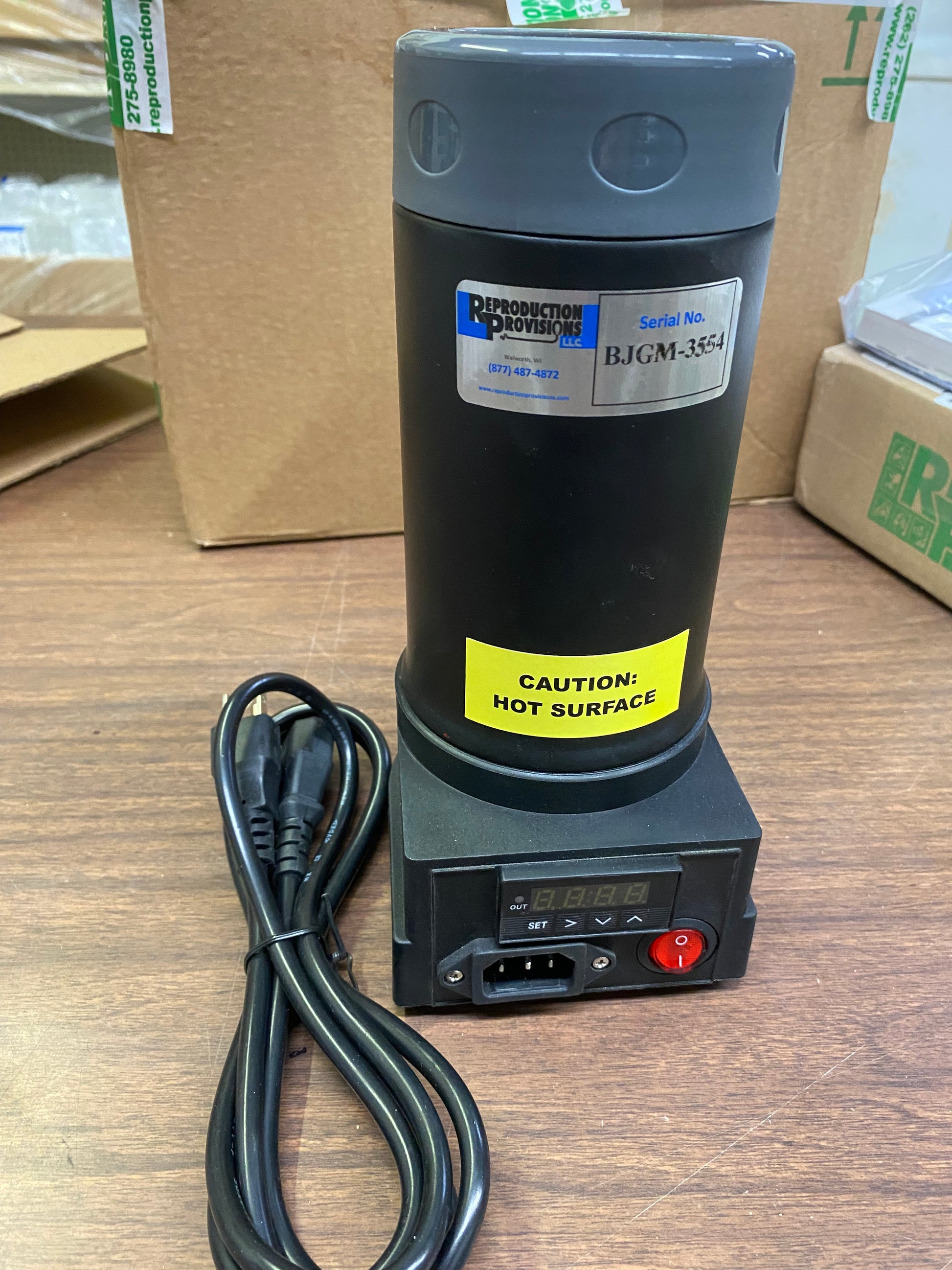 A black Digital Semen Thawing / Embryo Thawing Unit - 12V - 1023022 from Semen Tanks - Select Genetics sits on a wooden table, featuring a digital control base, 12V DC power cord, and a yellow CAUTION: HOT SURFACE sticker.