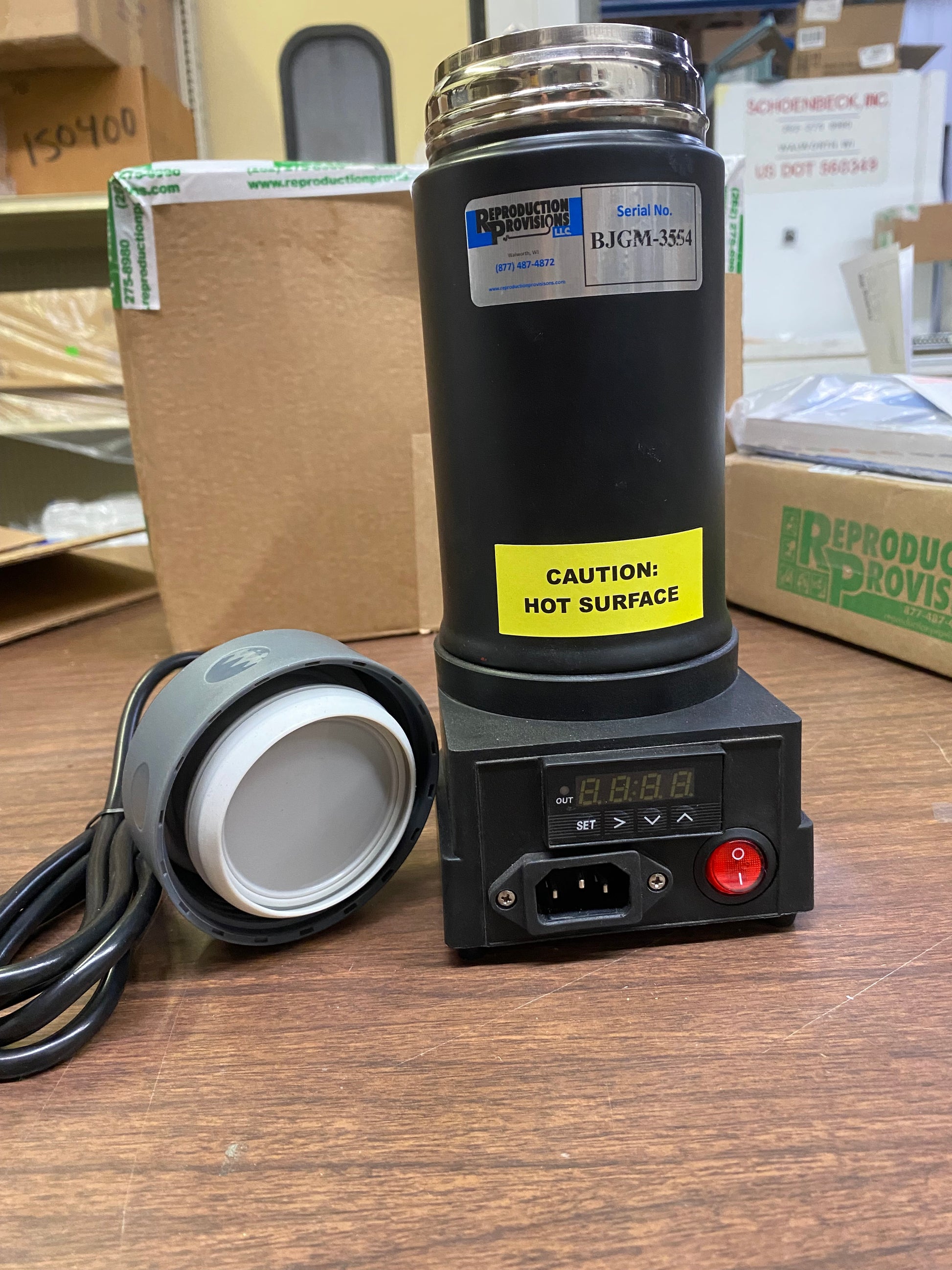 A black Semen Tanks - Select Genetics Digital Semen Thawing / Embryo Thawing Unit (12V, 1023022) sits on a digital heating base with a power cord and control panel, labeled Caution: Hot Surface, on a cluttered table with boxes in the background.