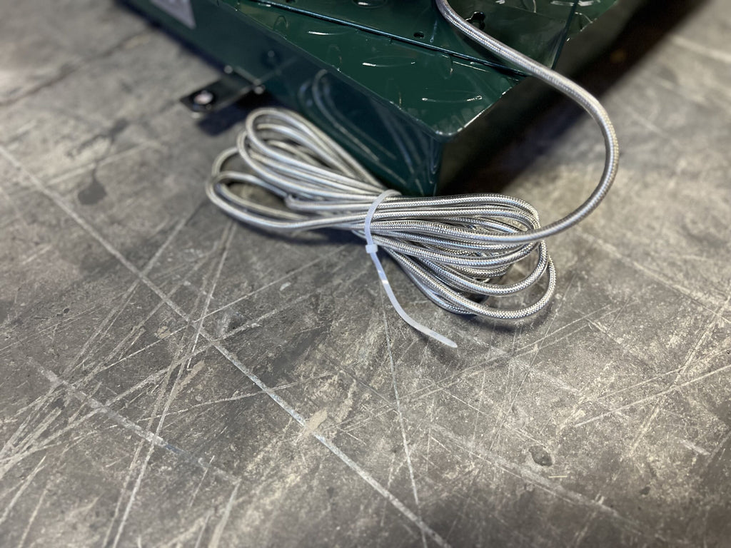 A coiled silver cable secured with a white twist tie sits on a scratched gray metal surface beside a green object, likely part of the Semen Tanks - Select Genetics CRYO-ALERT FLOOR SCALE Weight Indicator and Stack Light (110 lbs).