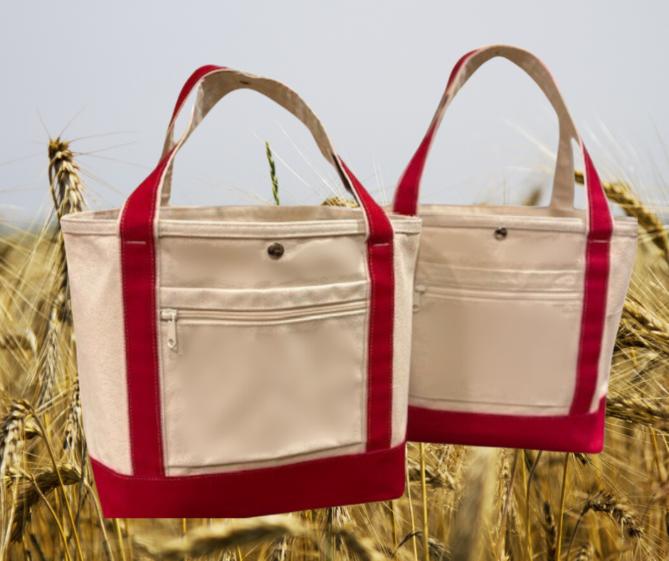 Two beige Semen Tanks - Select Genetics Totes for Everyday Use with red accents rest in a golden wheat field, crafted from durable duck canvas. Each tote offers roomy capacity, an exterior zip pocket, and your choice of 18 or 24 inch straps.
