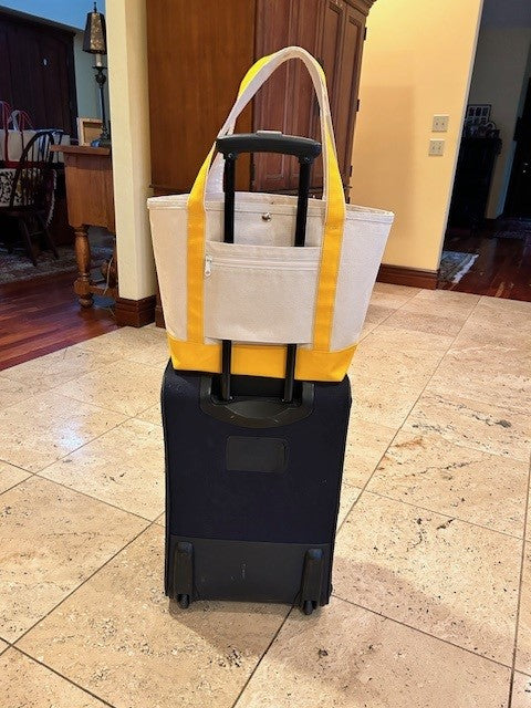 A Semen Tanks - Select Genetics Totes for Everyday Use bag with yellow accents and 24-inch straps rests atop a black rolling suitcase, handles looped over the suitcase handle, on a beige tiled floor in a home setting.