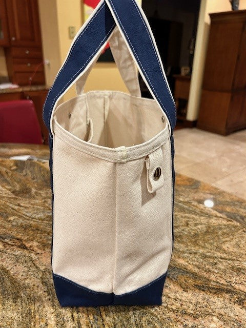 A Semen Tanks - Select Genetics Totes for Everyday Use with denim accents and 18 or 24 inch straps sits upright on a granite countertop. The cream tote features a snap button tab, blue handles and trim, with kitchen and wooden furniture in the background.