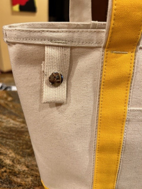 Close-up of the Semen Tanks - Select Genetics Totes for Everyday Use bag in beige duck canvas with yellow 24-inch straps and a fabric tab fastened by a brown marbled button. Blurry, warmly lit indoor background. Made in USA.