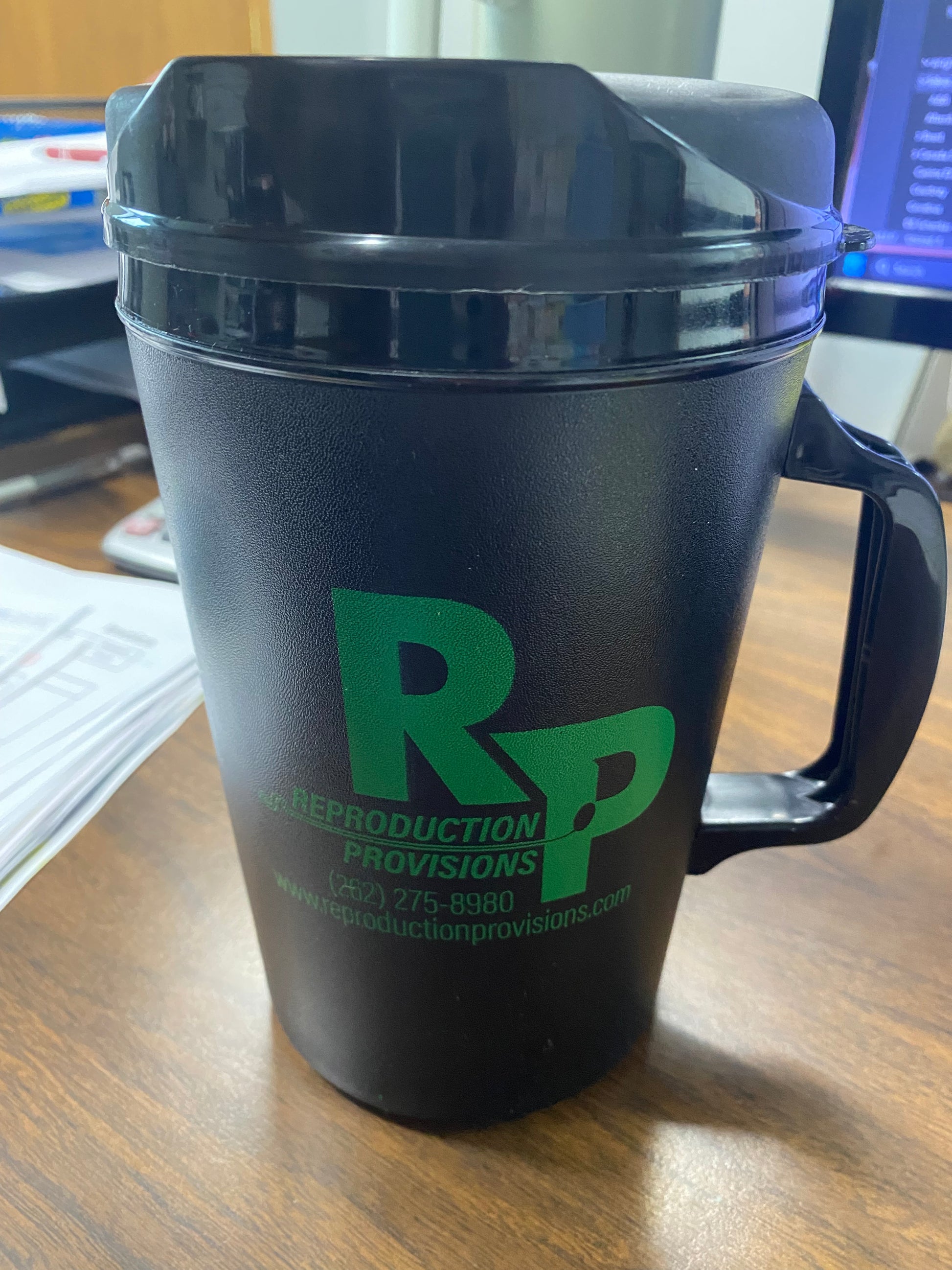 A large black RR Thermos with Thermometer, featuring a handle and lid, sits on a wooden desk. The mug displays a green RR logo with the product name below.