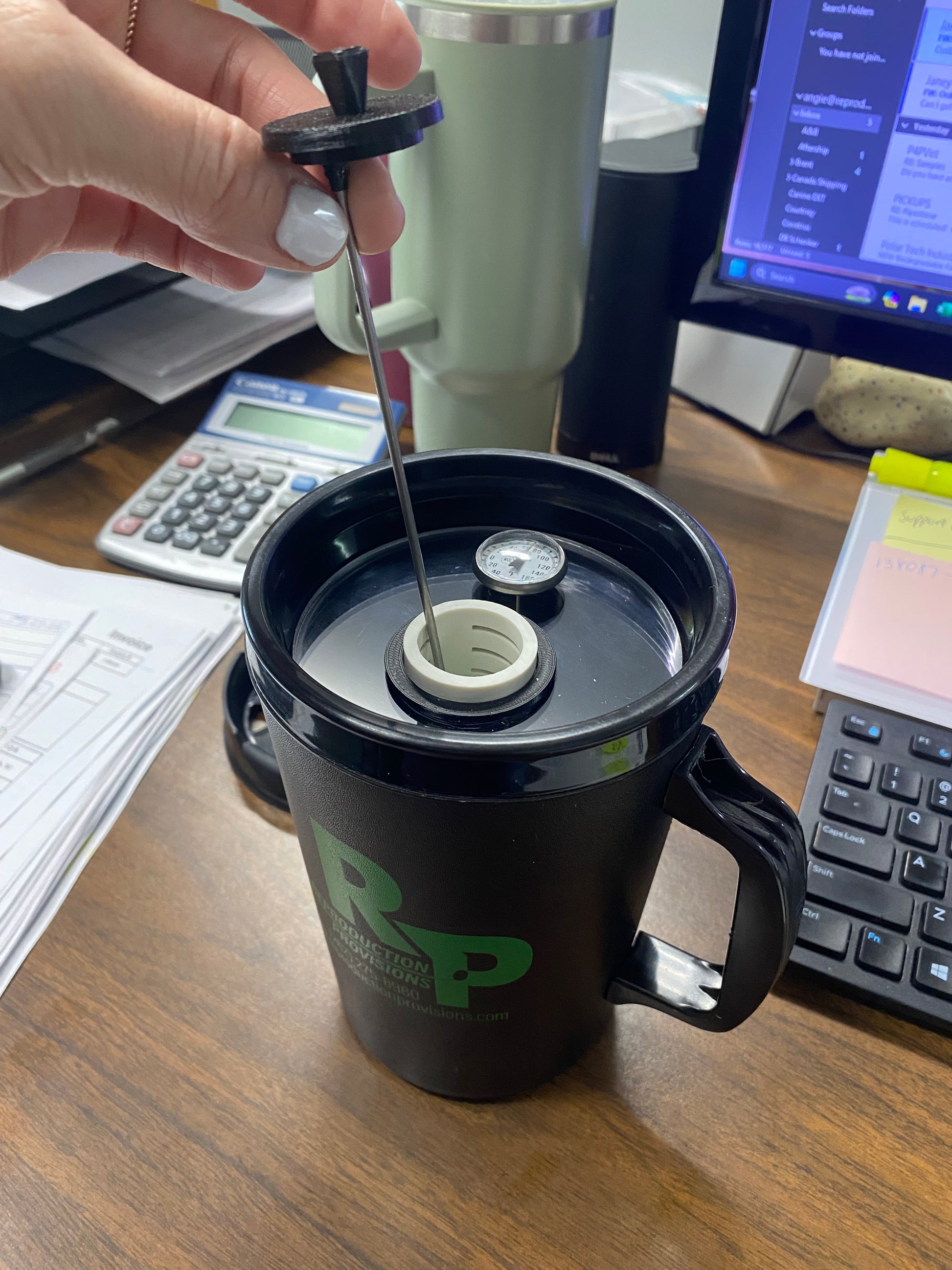 A hand lifts the lid and filter from a black RR Thermos with Thermometer, revealing its glass liner. On the desk are papers, a calculator, monitors, a banana, sticky notes, and the thermometer inserted in the thermos lid.